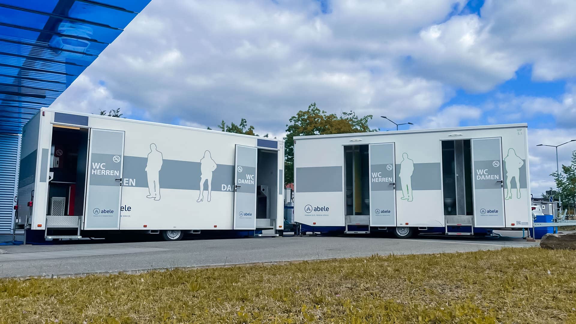 Mobile Toilettenwagen mit Beschriftungen für Herren und Damen auf einem Parkplatz unter bewölktem Himmel.