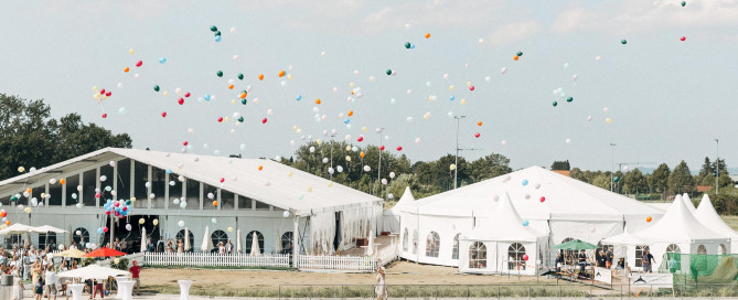 Menschen feiern bei weißen Zelten, viele bunte Luftballons steigen in den blauen Himmel, teils bewölkt und sonnig.