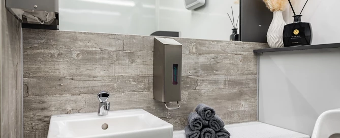 Waschbecken mit Wasserhahn, Seifenspender, graue Handtücher, Vase mit getrockneten Gräsern und Duftstäbchen auf Regal.