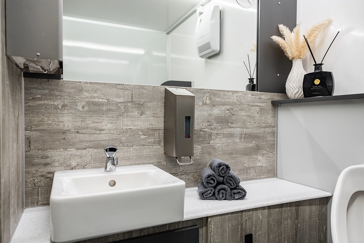 Waschbecken mit Wasserhahn, Seifenspender, graue Handtücher, Vase mit getrockneten Gräsern und Duftstäbchen auf Regal.