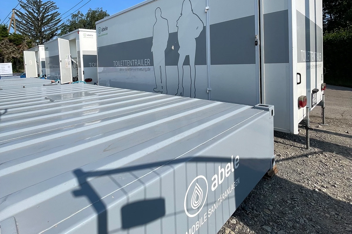 Mobile Sanitäranlagen auf einem Schotterplatz; mehrere Toilettentrailer der Marke abele in einer Reihe mit offenen Türen.