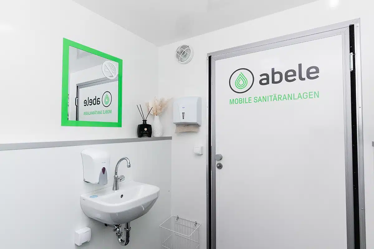 Toilettencontainer Handicap 5 Moderner Sanitärraum mit Waschbecken, Spiegel und Handtuchspender. Tür mit Abele-Logo für mobile Sanitäreinheiten.