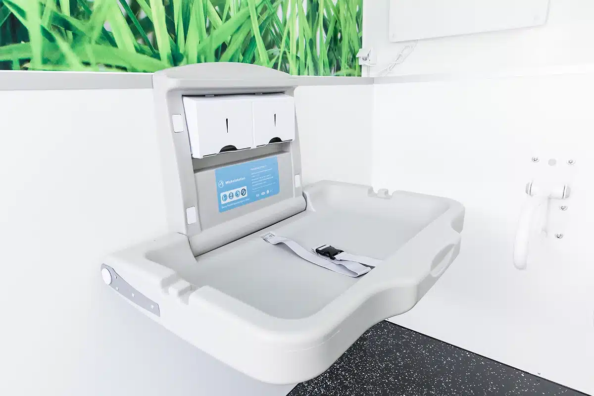Toilettencontainer Handicap 4 Wickeltisch mit Gurt in modernem, weißem Raum. Grüne Pflanzenwand und Haltegriff sind sichtbar.