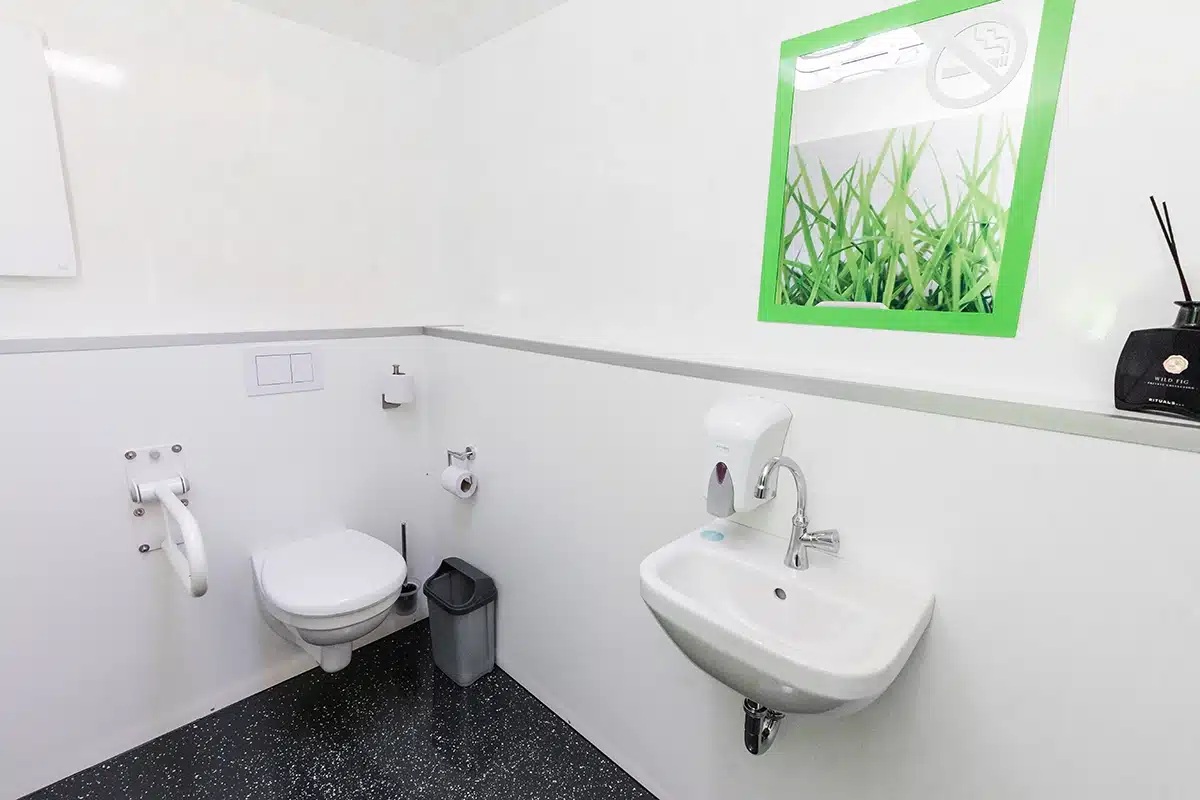 Toilettencontainer Handicap 7 Barrierefreies Bad mit Wandtoilette, Waschbecken, Haltegriff, Papierrollenhalter, Mülleimer und grün gerahmtem Spiegel.