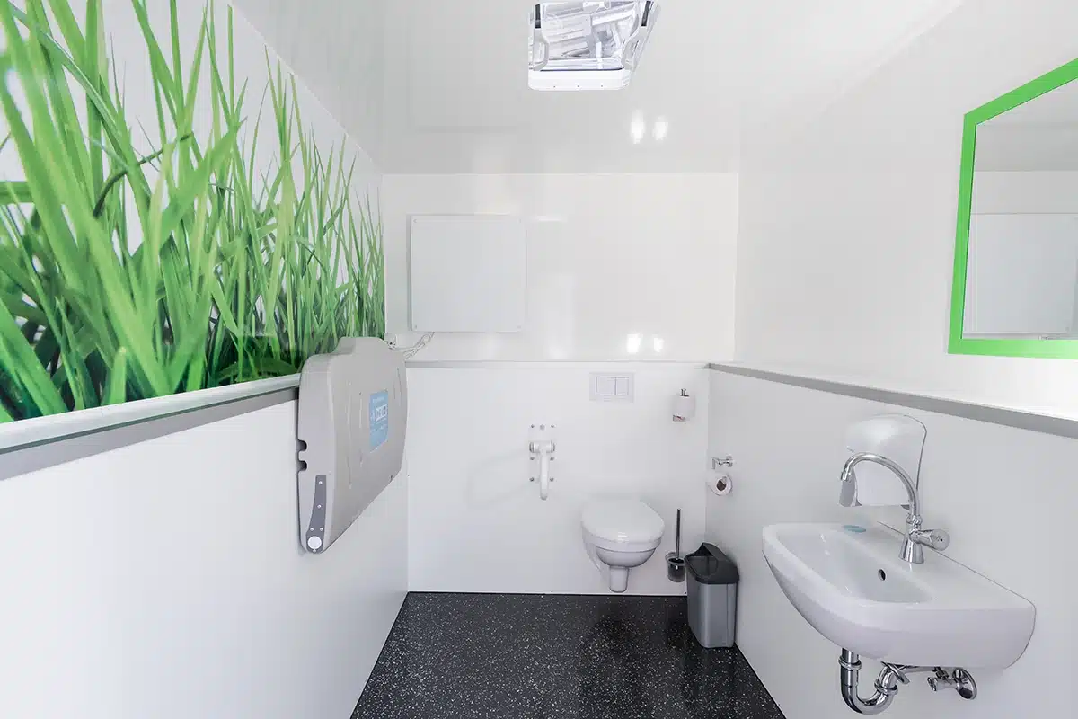 Toilettencontainer Handicap 6 Barrierefreies WC mit Wickeltisch, Waschbecken, grünem Wandbild von Gras und grellem Spiegelrahmen auf schwarzem Boden.