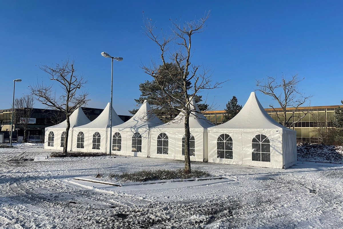 Weiße Zelte mit spitzen Dächern stehen nebeneinander auf verschneitem Boden, umgeben von kahlen Bäumen bei klarem Himmel.