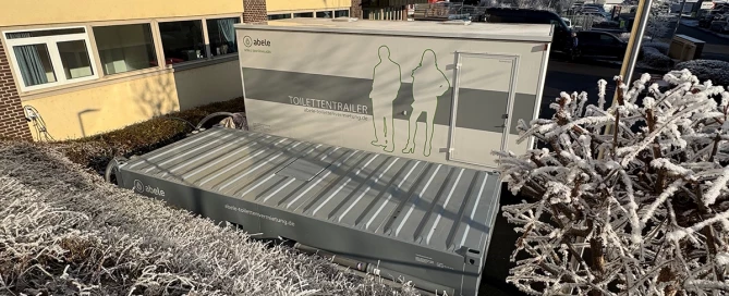 Toilettencontainer neben einem Gebäude, umgeben von gefrorenen Büschen und Bäumen im Winter.