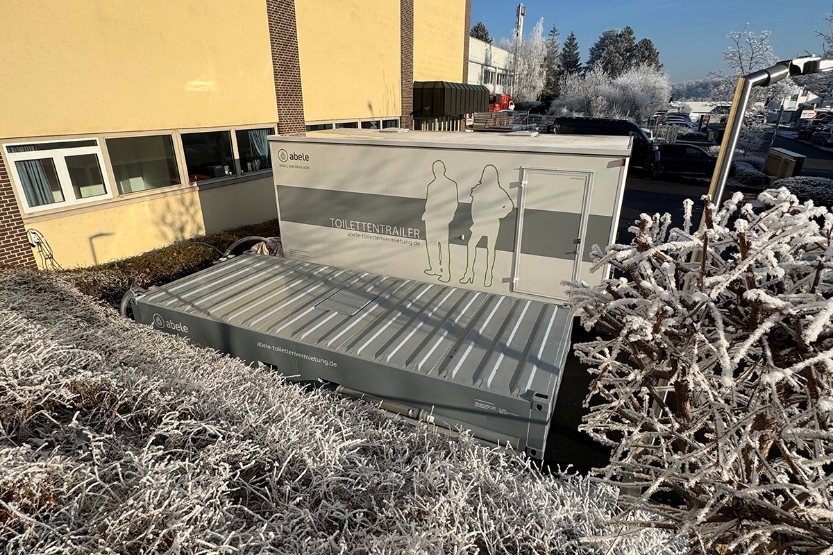 Toilettencontainer neben einem Gebäude, umgeben von gefrorenen Büschen und Bäumen im Winter.