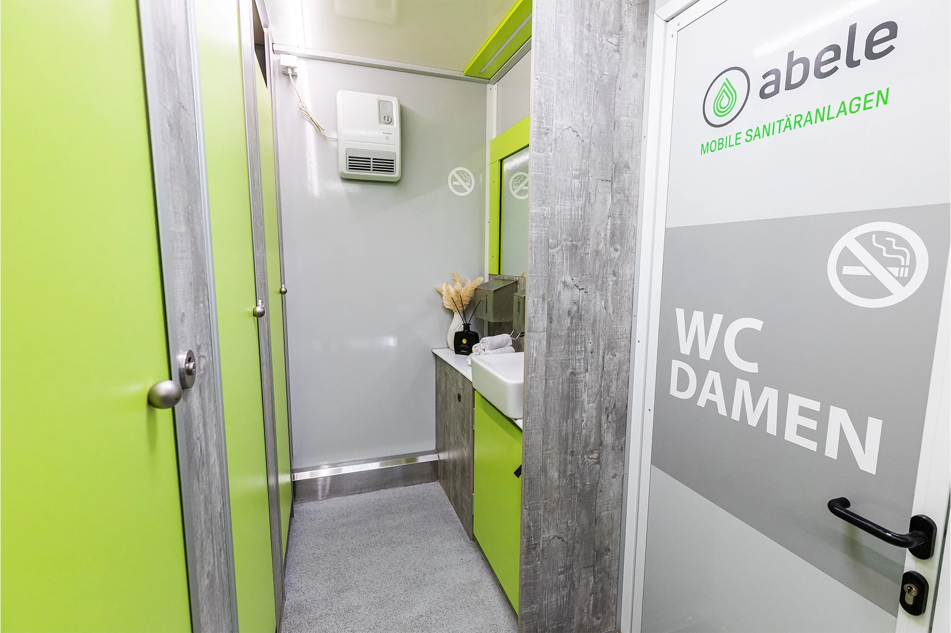 Luxus Toilettenwagen Modell Exklusiv – GoGreen 6 Moderne Damentoilette mit grünen Türen, Waschbecken, Spiegel und einem No-Smoking-Zeichen an der Tür.