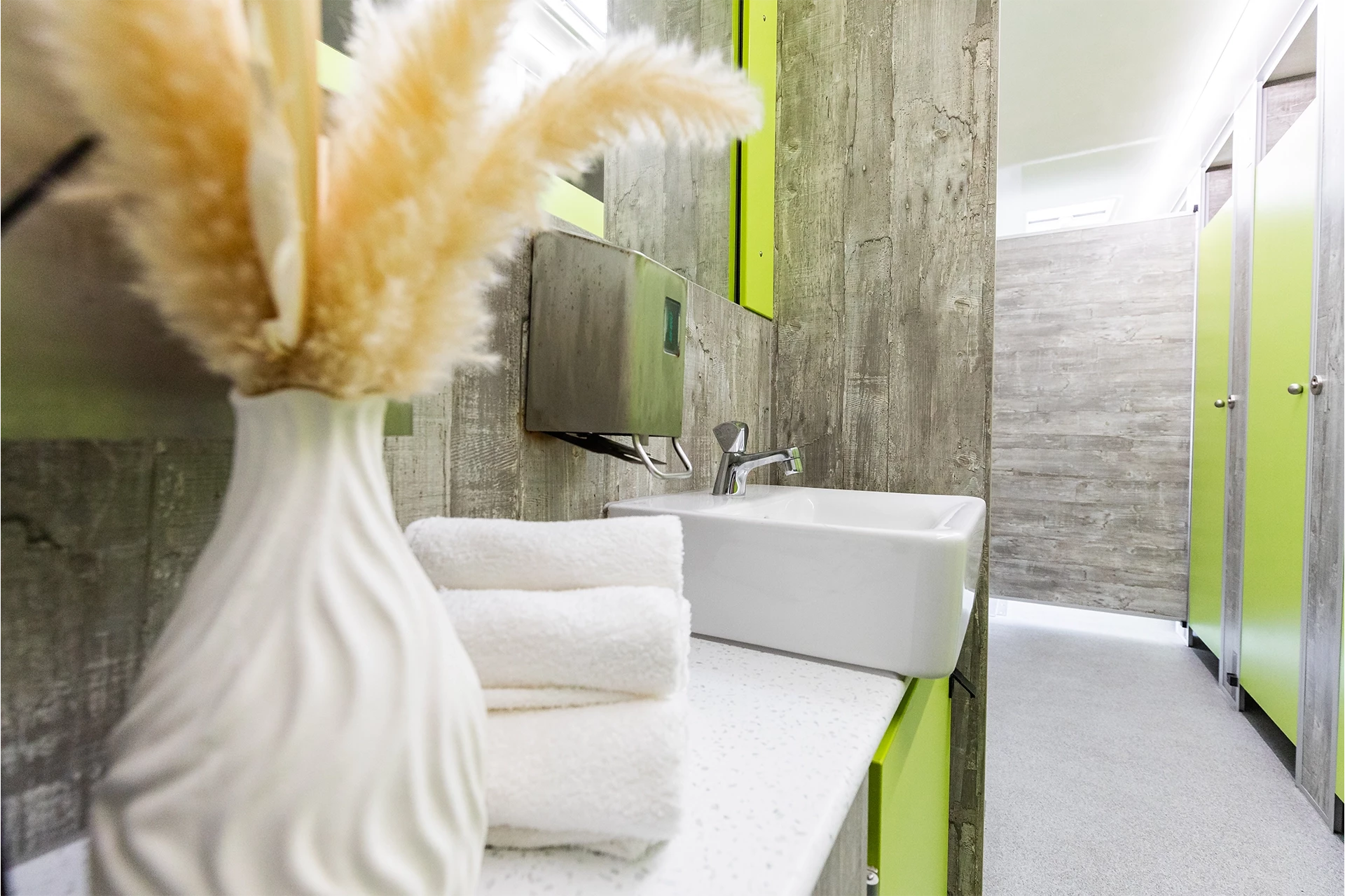 Luxus Toilettenwagen Modell Exklusiv – GoGreen 7 Modernes, graues Bad mit weißem Waschbecken, grünen Akzenten, Handtüchern und Pampasgras in der Vase.