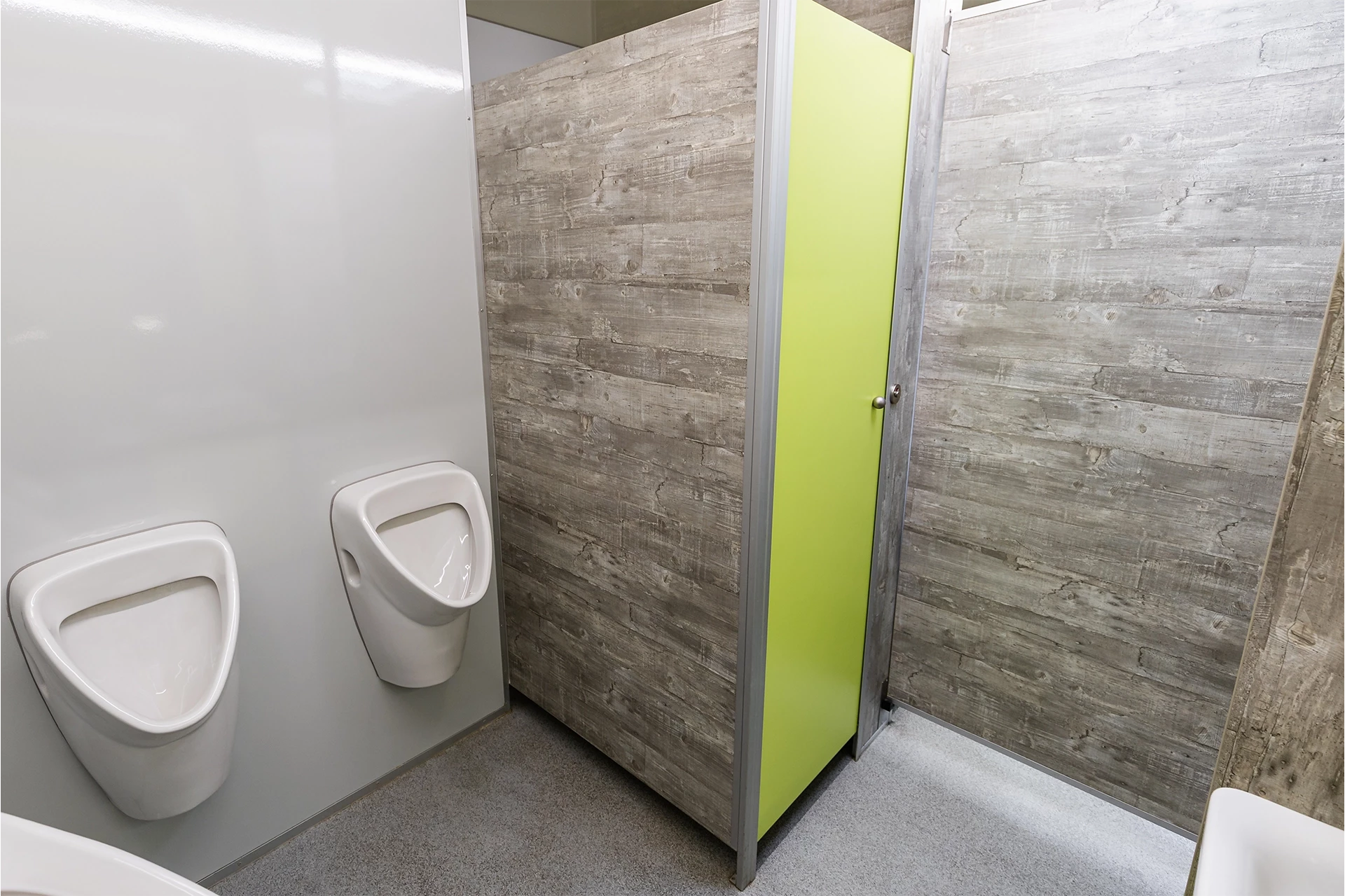 Luxus Toilettenwagen Modell Exklusiv – GoGreen 3 Zwei Urinale an der Wand neben einer Toilettenkabine mit grünem Türblatt in einem modern gefliesten Bad.