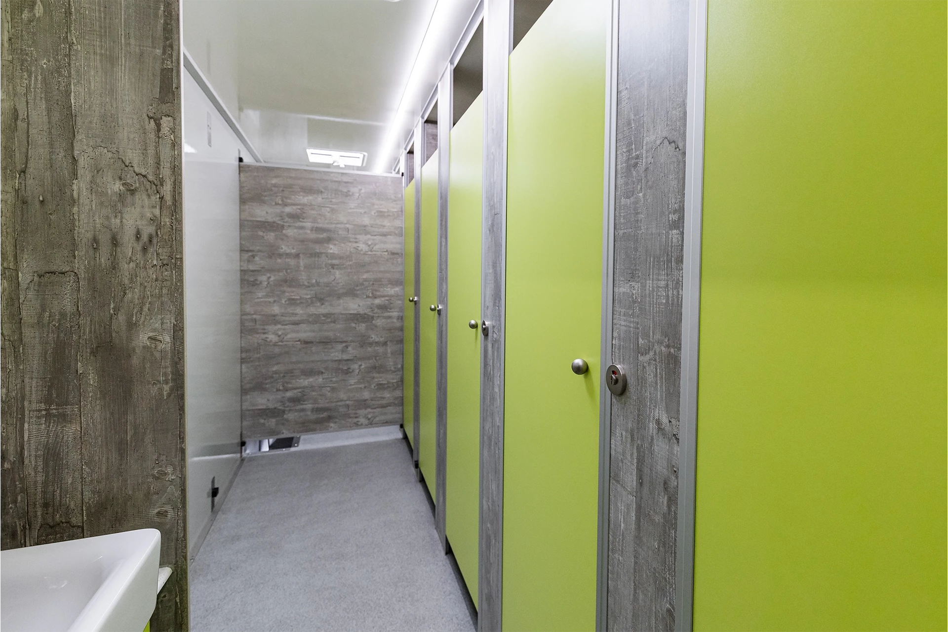 Luxus Toilettenwagen Modell Exklusiv – GoGreen 4 Mehrere Toilettenkabinen mit grünen Türen und grauen Wänden in einem sauberen, modernen Badezimmer. Links ist ein Waschbecken.