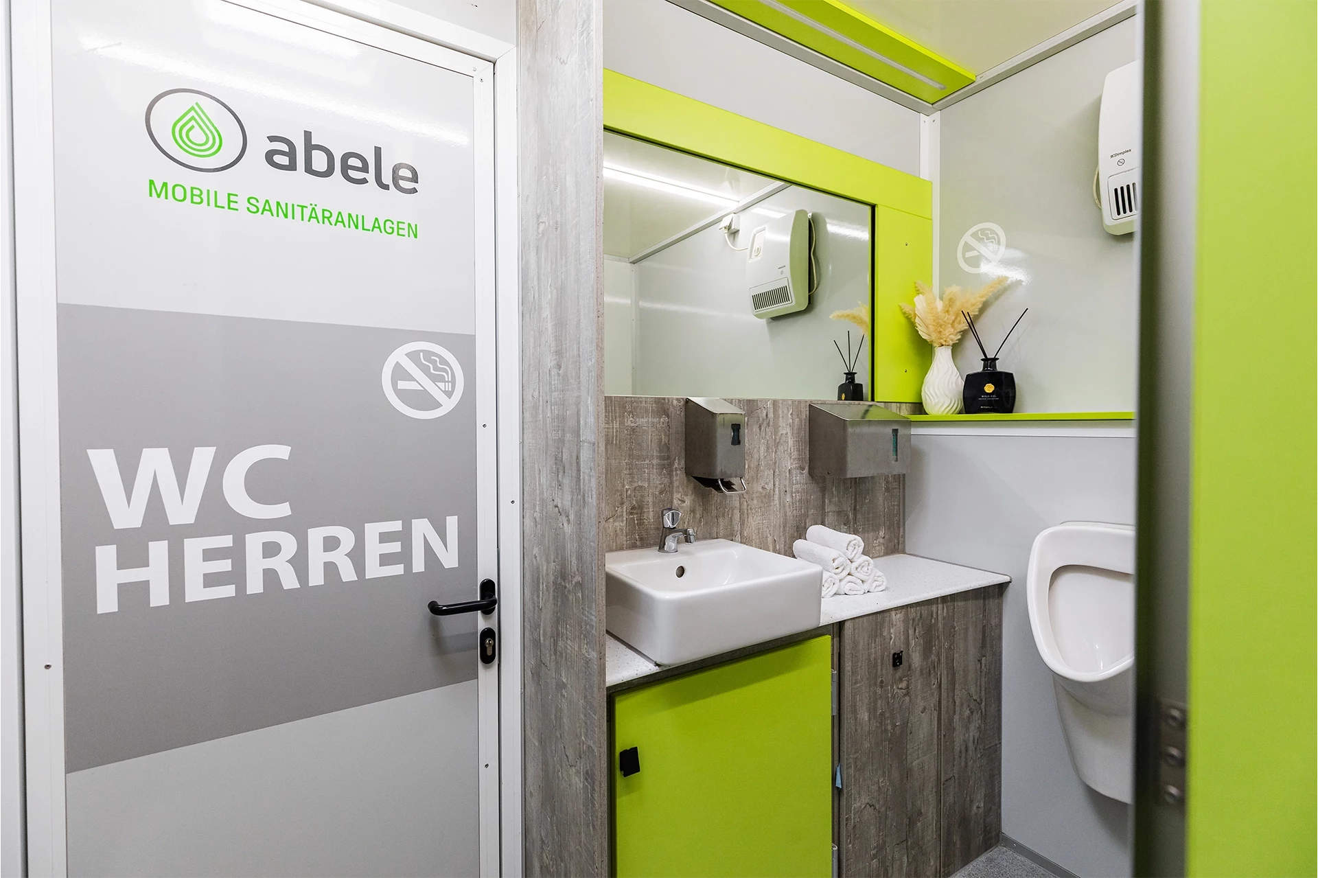 Luxus Toilettenwagen Modell Exklusiv – GoGreen 13 Grüne mobile Herren-Toilette mit Waschbecken, Spiegel, Handtuchspender, Pissoir und Deko in hellem, modernen Design.