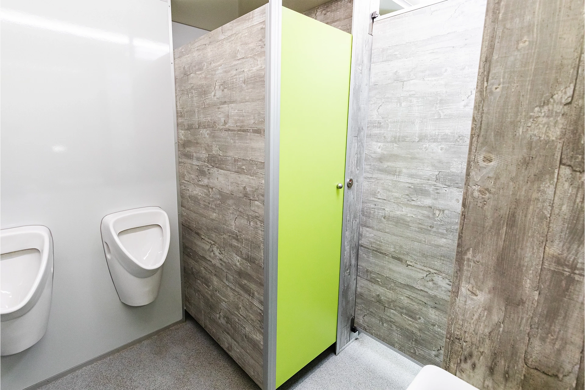 Luxus Toilettenwagen Modell Exklusiv – GoGreen 11 Öffentliche Toiletten mit Urinalen linkerhand und Kabine mit grüner Tür rechts, umgeben von grauen Trennwänden.
