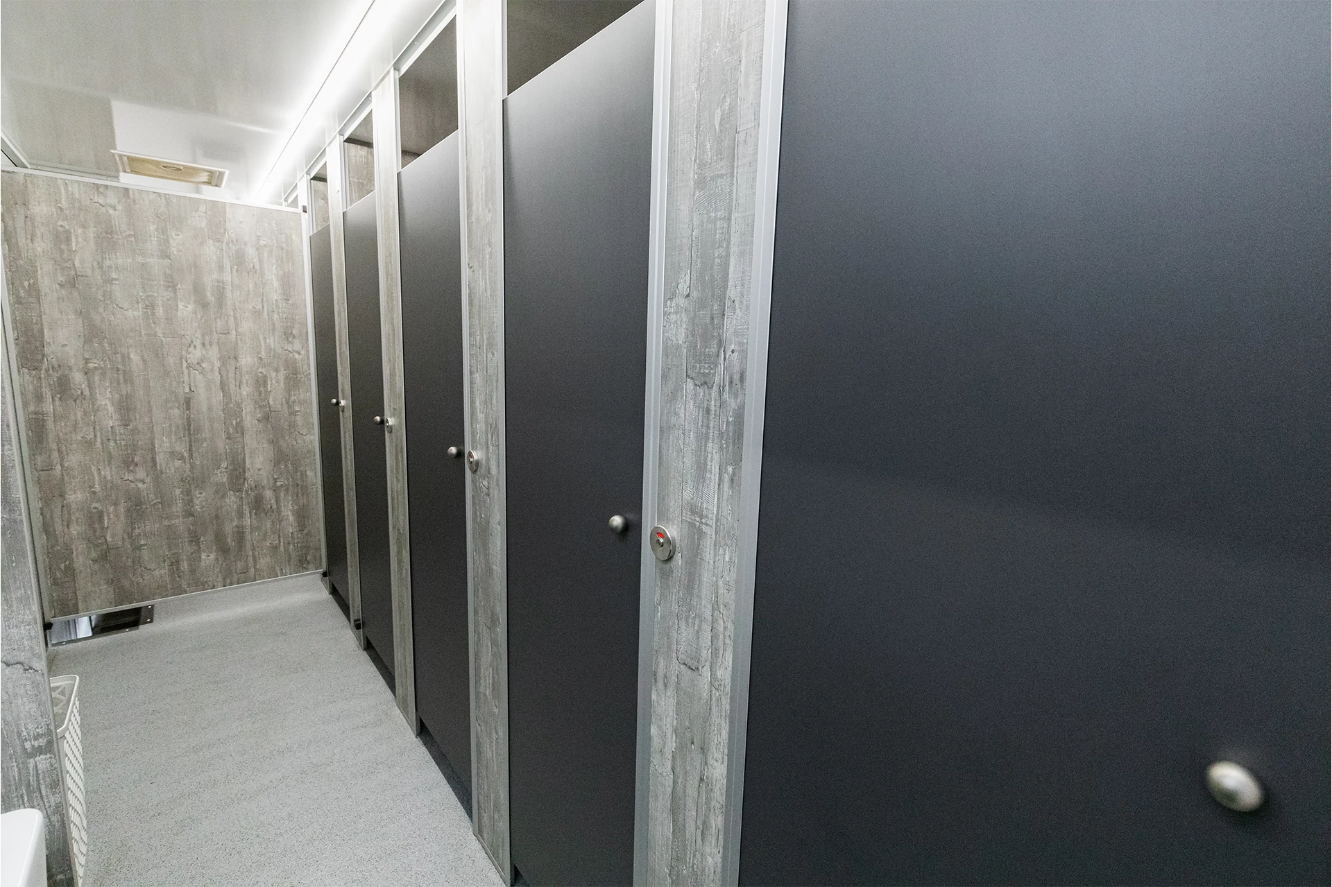 Toilettenwagen Modell Exklusiv – GoGrey 4 Reihe grauer Toilettenkabinen mit abgeschlossenen Türen in einem öffentlichen Waschraum. Dezente Holzmaserung an den Wänden.