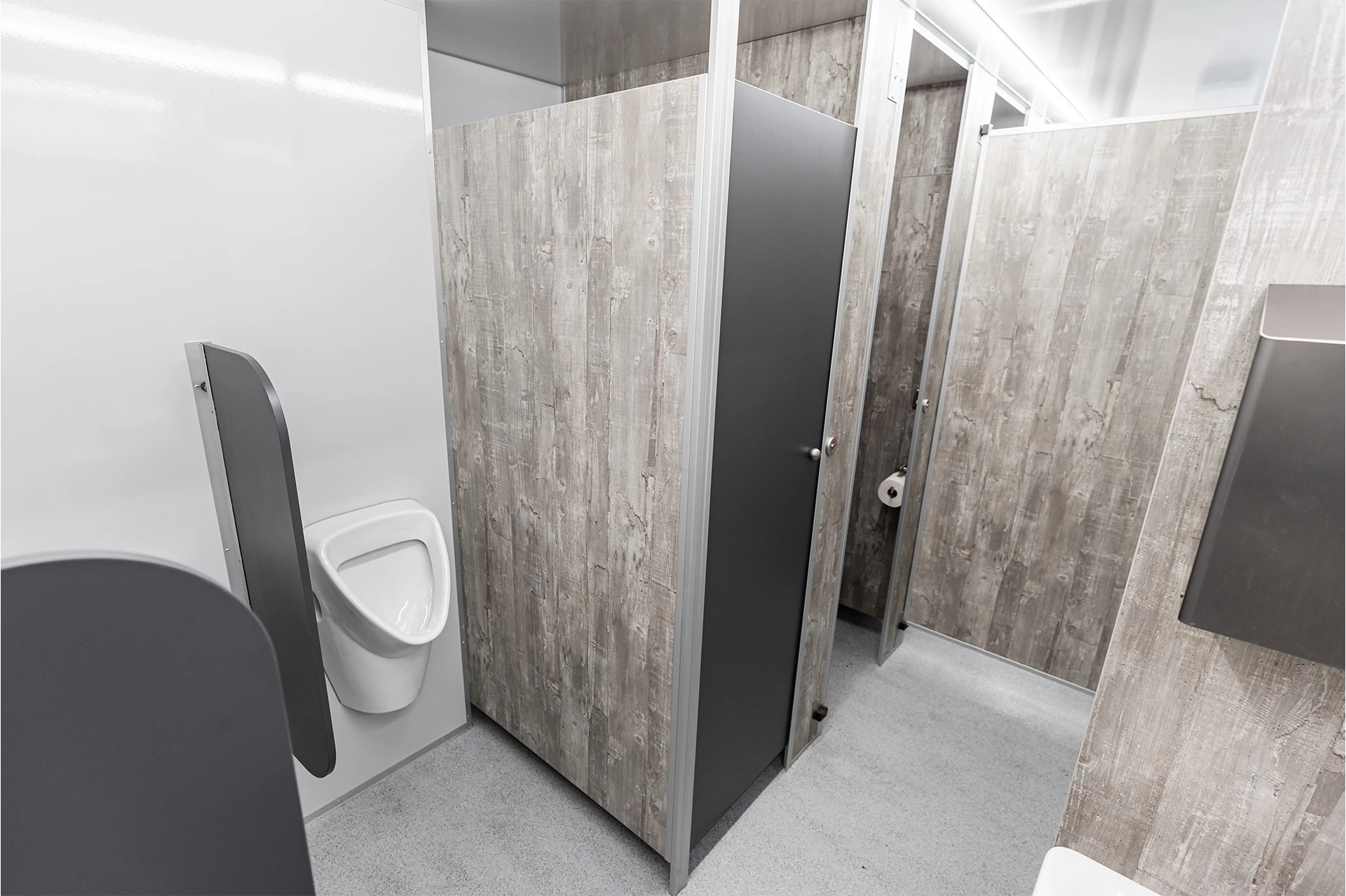 Toilettenwagen Modell Exklusiv – GoGrey 2 Helle öffentliche Toiletten mit Trennwänden aus Holzoptik; sichtbar sind ein Urinal und eine verschlossene Kabine.