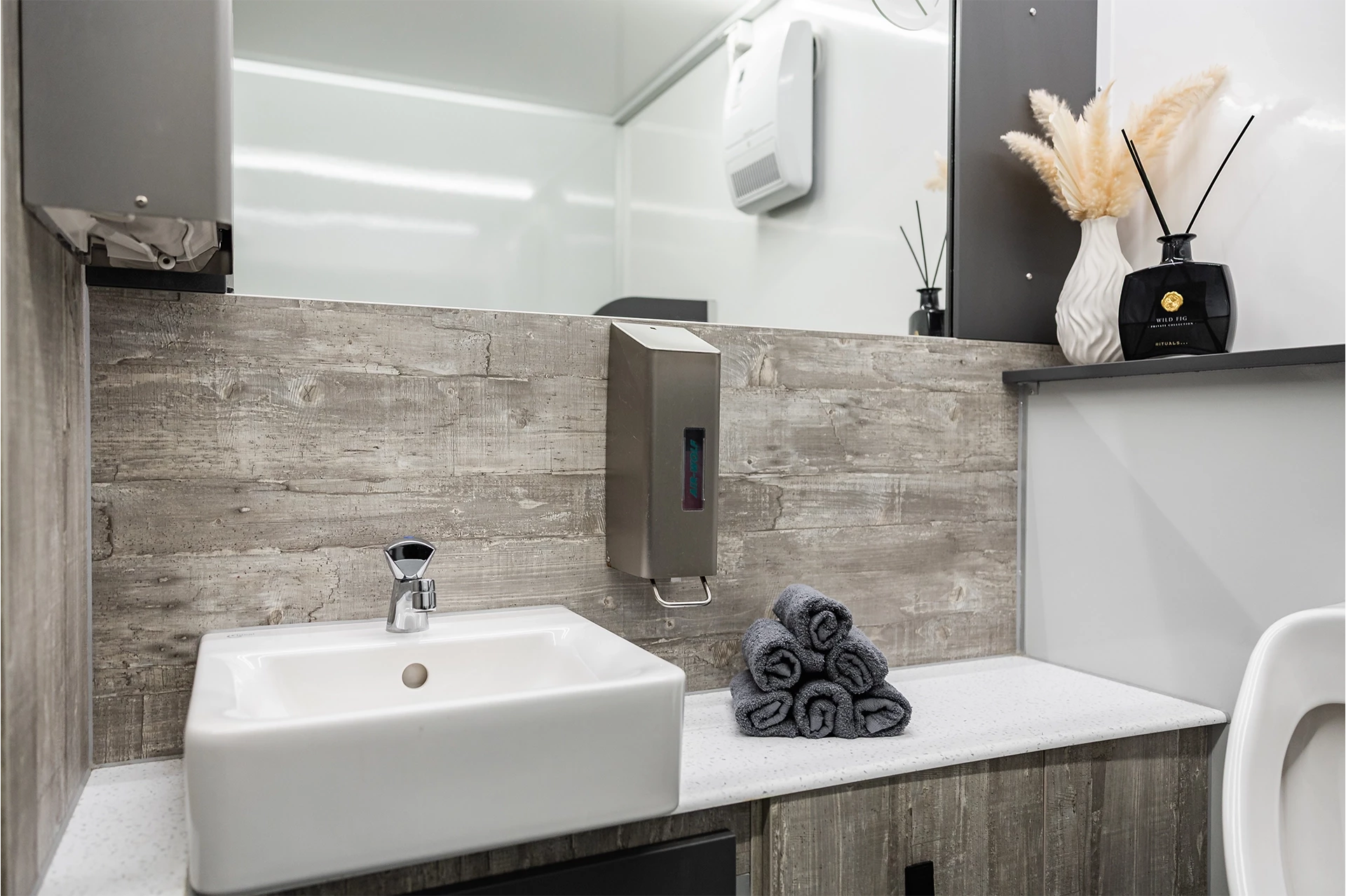 Toilettenwagen Modell Exklusiv – GoGrey 10 Modernes Badezimmer mit Waschtisch, Spiegel, Seifenspender und grauen Handtüchern auf weißer Ablage vor Holzwand.