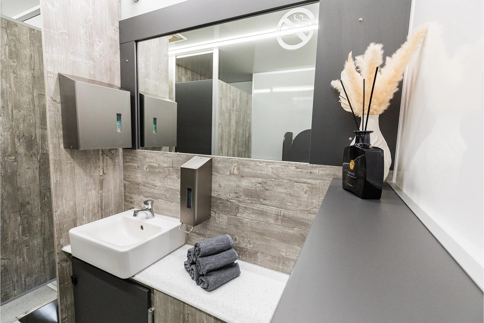 Toilettenwagen Modell Exklusiv – GoGrey 8 Modernes Badezimmer mit Waschtisch, Spiegel, Handtuchstapel, Seifenspender und Deko-Gräsern in schwarzer Vase.