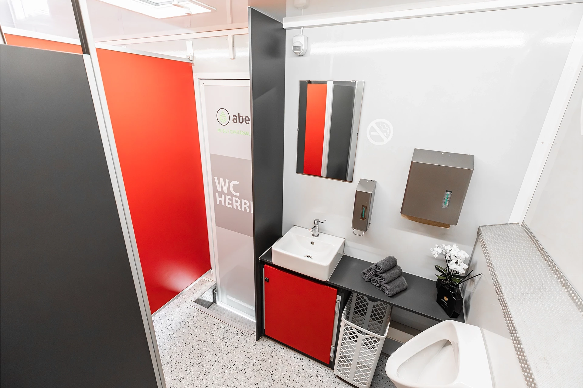 Toilettenwagen Modell Exklusiv – Mix 5/1 8 Moderne, saubere Herrentoilette mit roten Akzenten, Waschbecken, Spiegel, Papierhandtuchspender und Orchidee auf der Ablage.