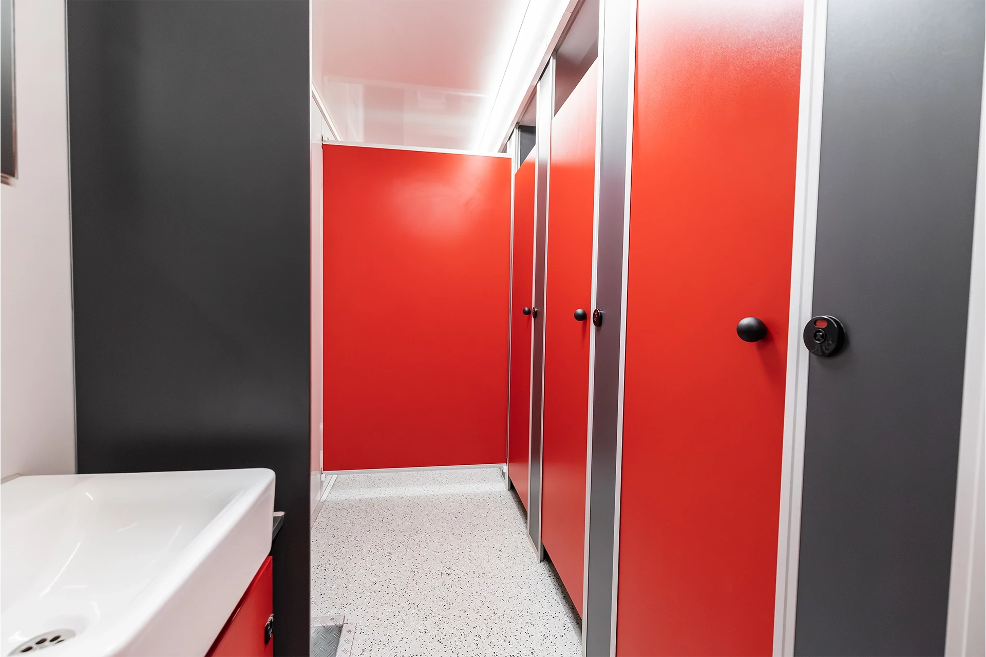 Toilettenwagen Modell Exklusiv – Mix 5/1 2 Rote WC-Kabinentüren in einem modernen Waschraum mit Waschbecken und grauen Wänden auf hellem, gesprenkeltem Boden.