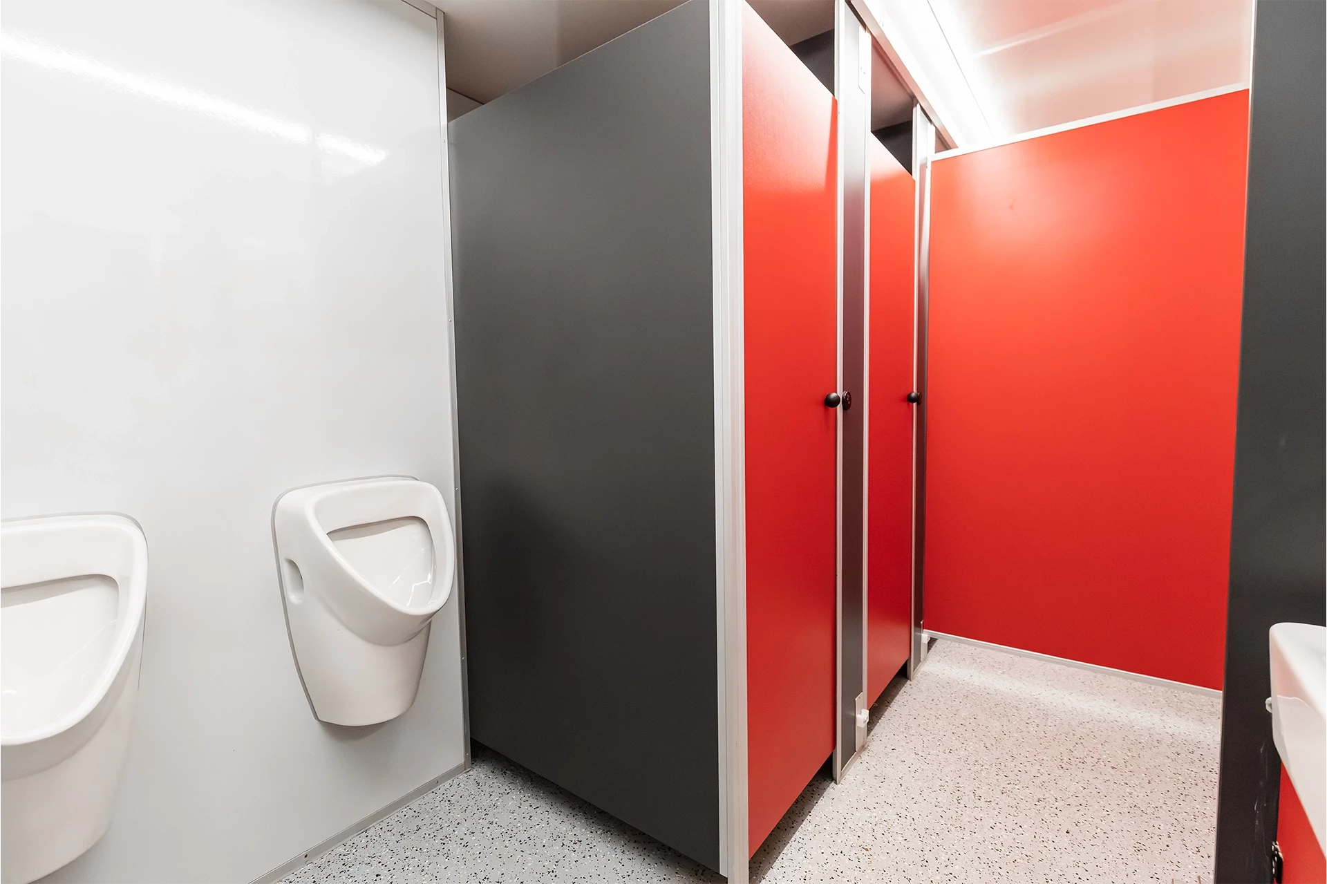 Toilettenwagen Modell Exklusiv – Mix 5/1 7 Öffentliche Toilette mit roten Kabinentüren und zwei Urinalen an einer weißen Wand, grauer Bodenbelag.