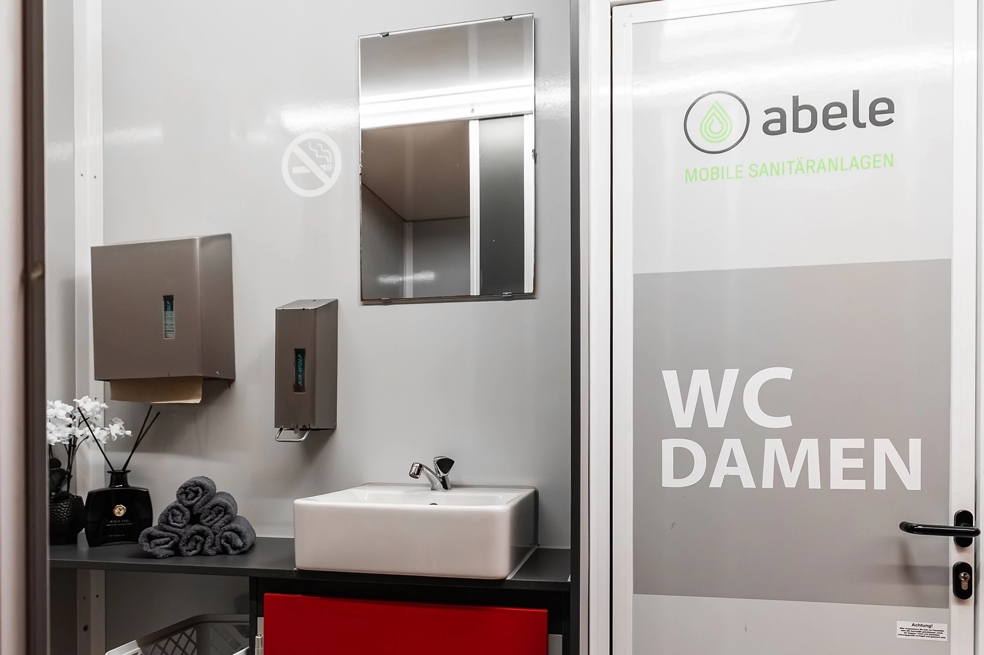 Toilettenwagen Modell Exklusiv – Mix 5/1 6 Damen-WC mit Waschbecken, Spiegel, Handtuchspendern und Deko, keine Rauchen-Schild, von Abele Mobile Sanitäranlagen.