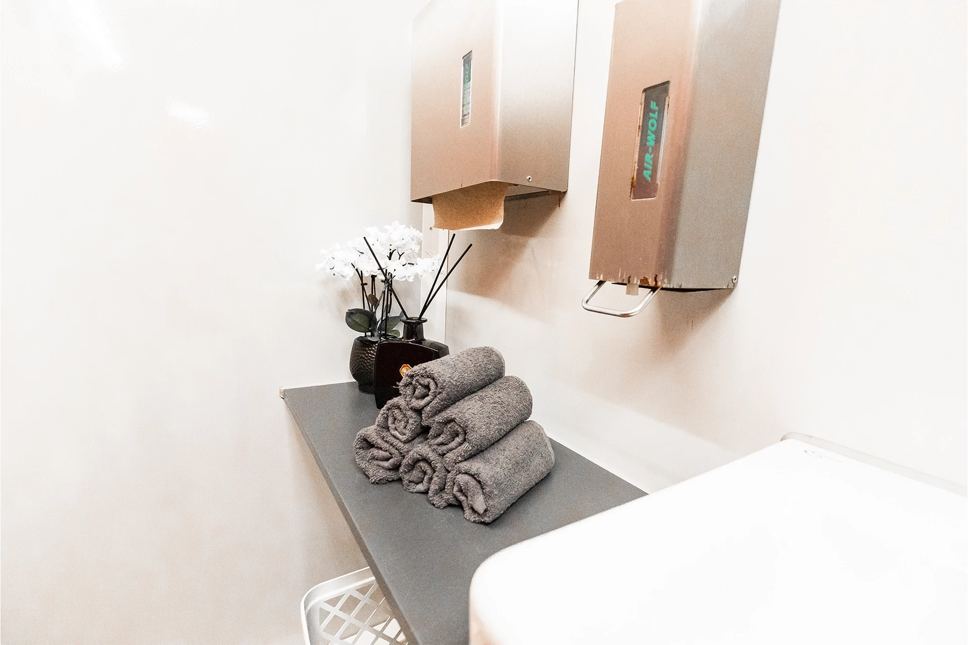 Toilettenwagen Modell Exklusiv – Mix 5/1 5 Graue Handtücher auf Tisch, daneben Vase mit weißen Blumen und Duftstäbchen. An der Wand zwei Papierhandtuchspender.