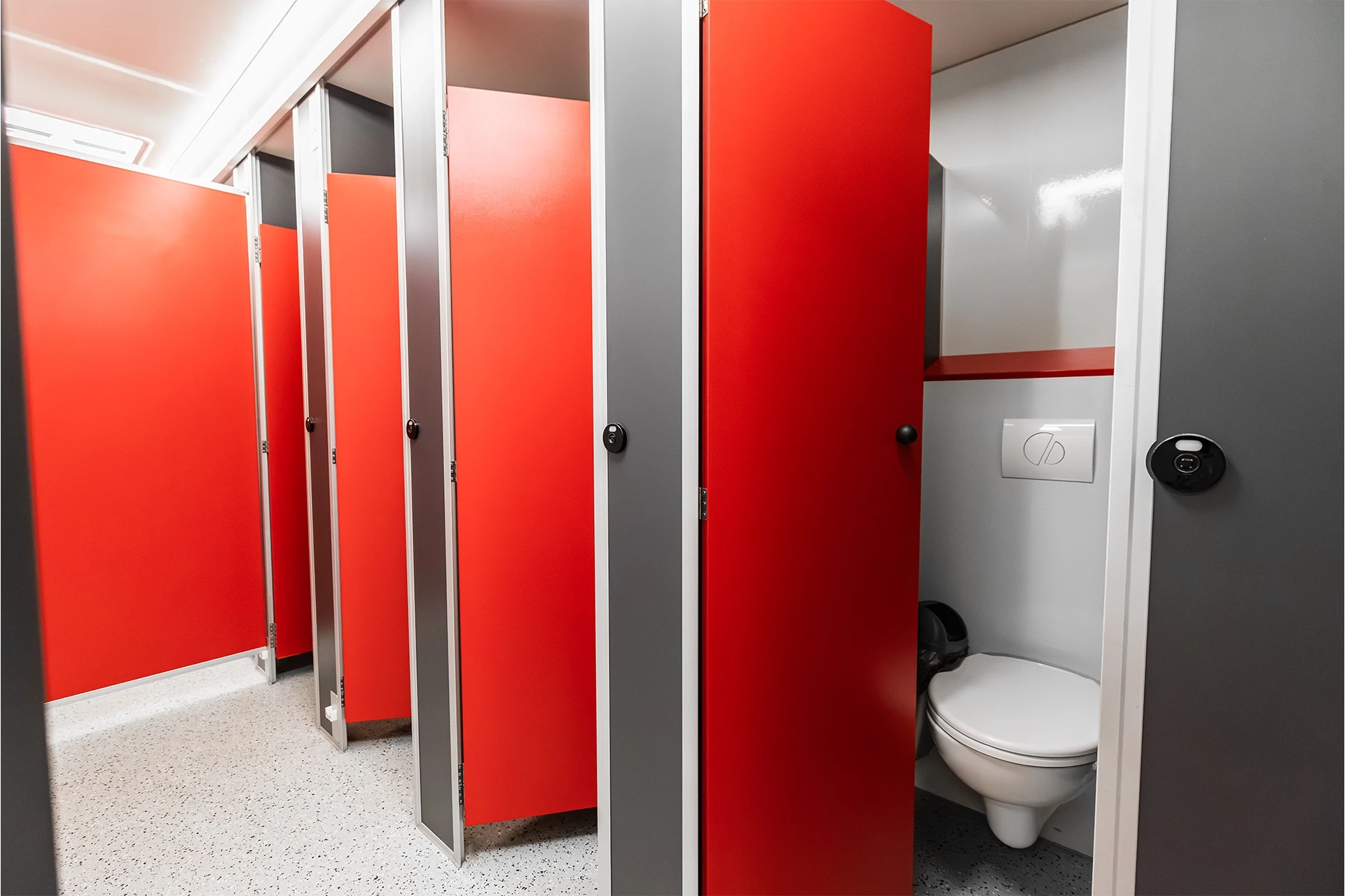 Toilettenwagen Modell Exklusiv – Mix 5/1 3 Rote Toilettenkabinen, teils geschlossen, eine zeigt eine weiße Toilette und Spülknopf. Grauer, gefliester Boden.