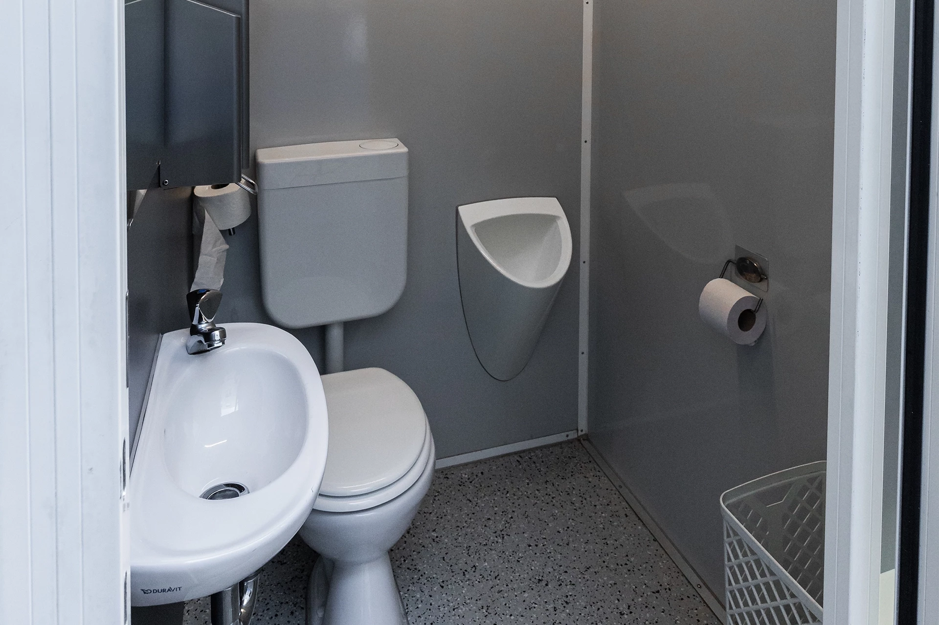 Toilettenwagen Modell Exklusiv – Handicap +2 9 Kleine, graue Toilette mit WC, Urinal, Waschbecken und Toilettenpapier. Boden mit grauem Muster.