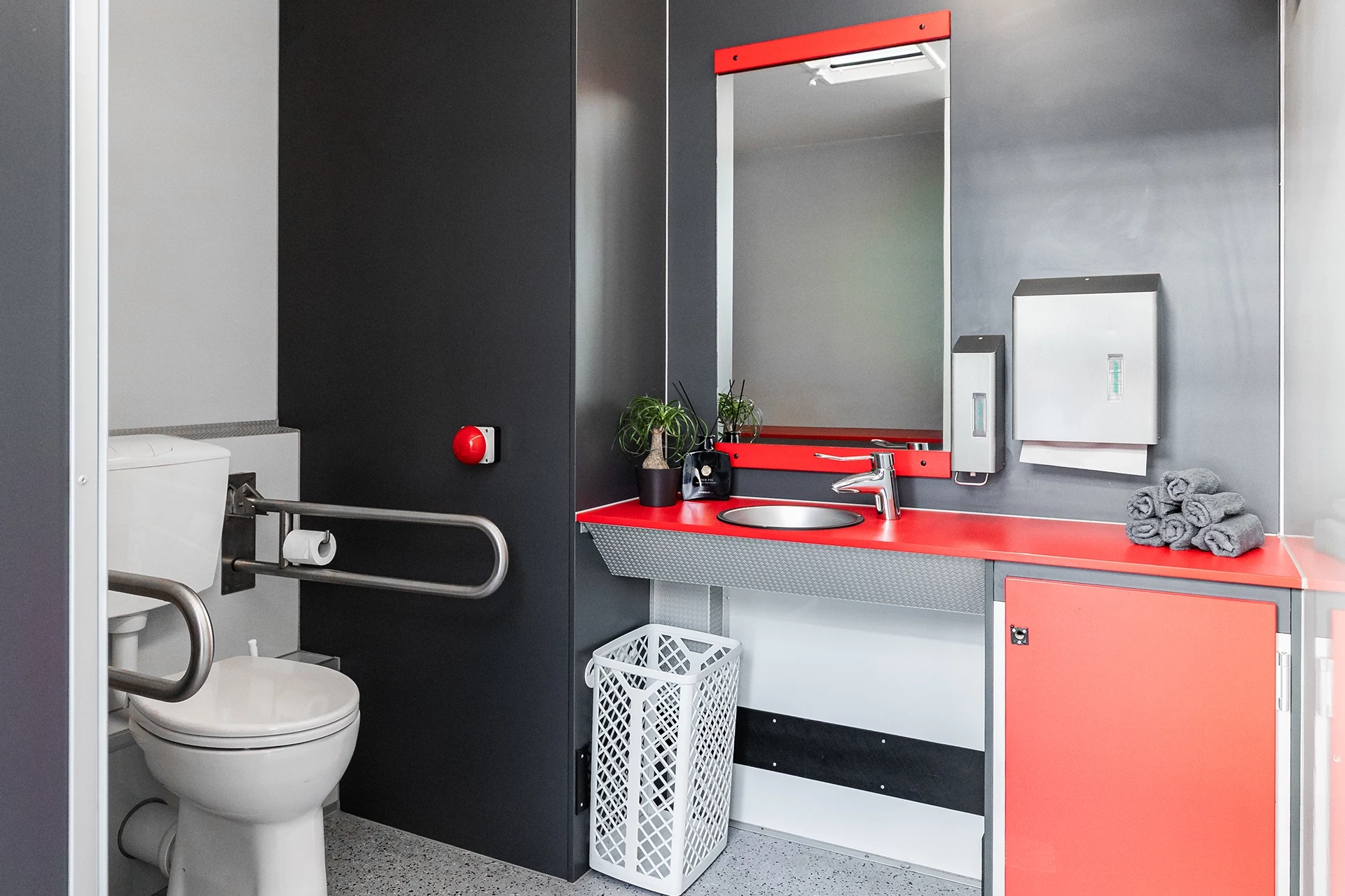 Toilettenwagen Modell Exklusiv – Handicap +2 5 Barrierefreies Badezimmer mit Haltegriffen am WC, rotem Waschbecken, Spiegel, Pflanzen, Handtuchhalter und Notrufknopf.