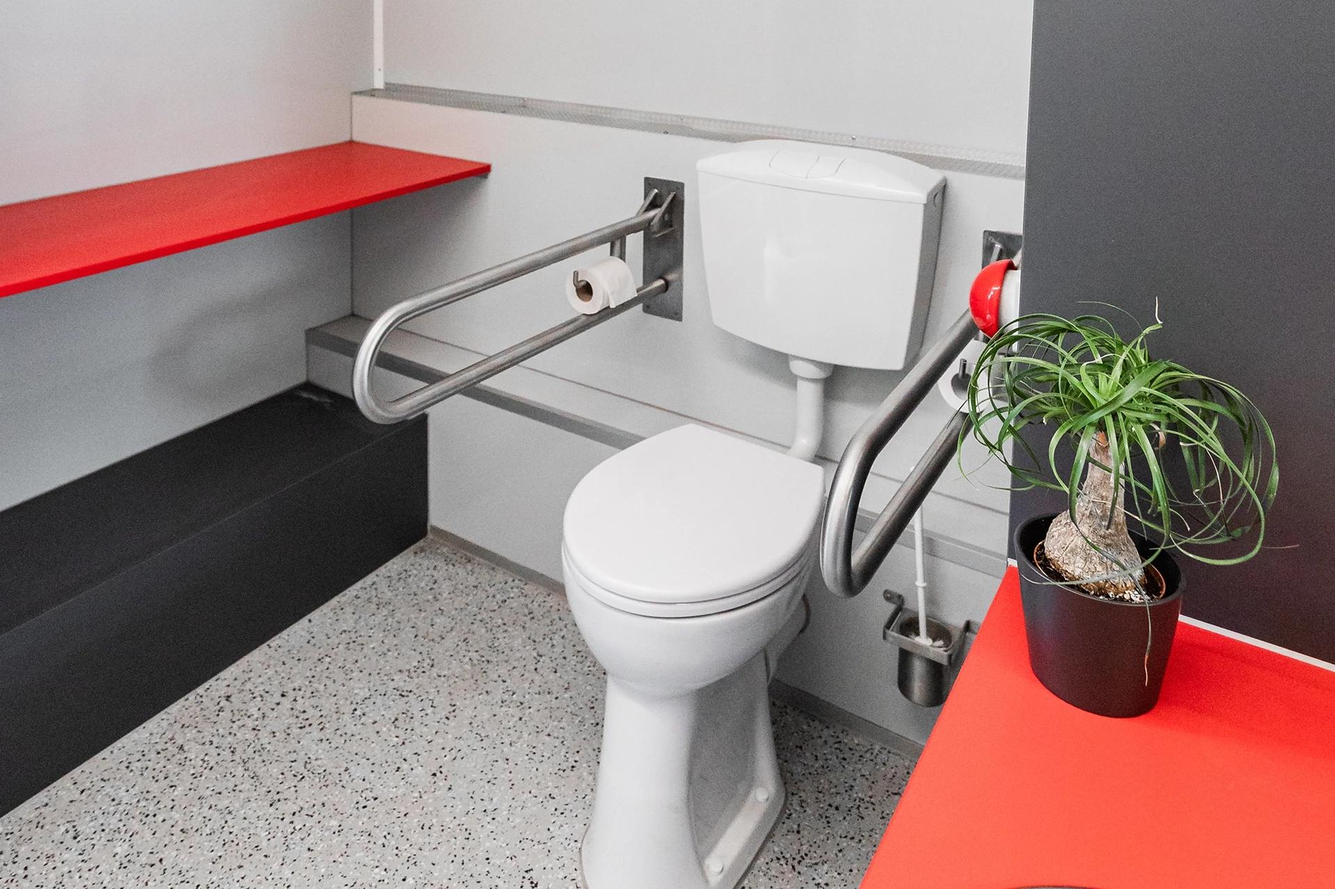 Toilettenwagen Modell Exklusiv – Handicap +2 4 Barrierefreie Toilette mit Haltegriffen, Papierhalter und rotem Regal. Eine Pflanze steht auf dem Regal neben der Toilette.