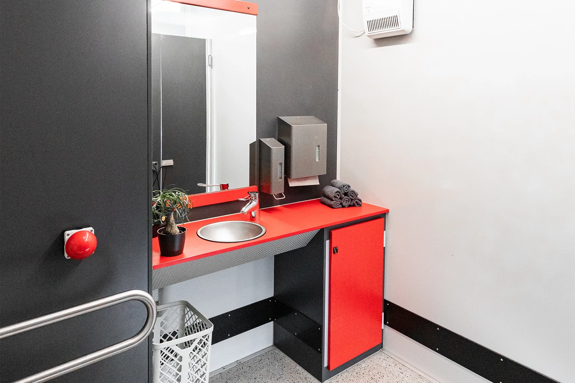 Toilettenwagen Modell Exklusiv – Handicap +2 2 Barrierefreies Badezimmer: Waschbecken mit Spiegel, rotem Notknopf, Handtüchern und dekorativer Pflanze auf rotem Unterschrank.
