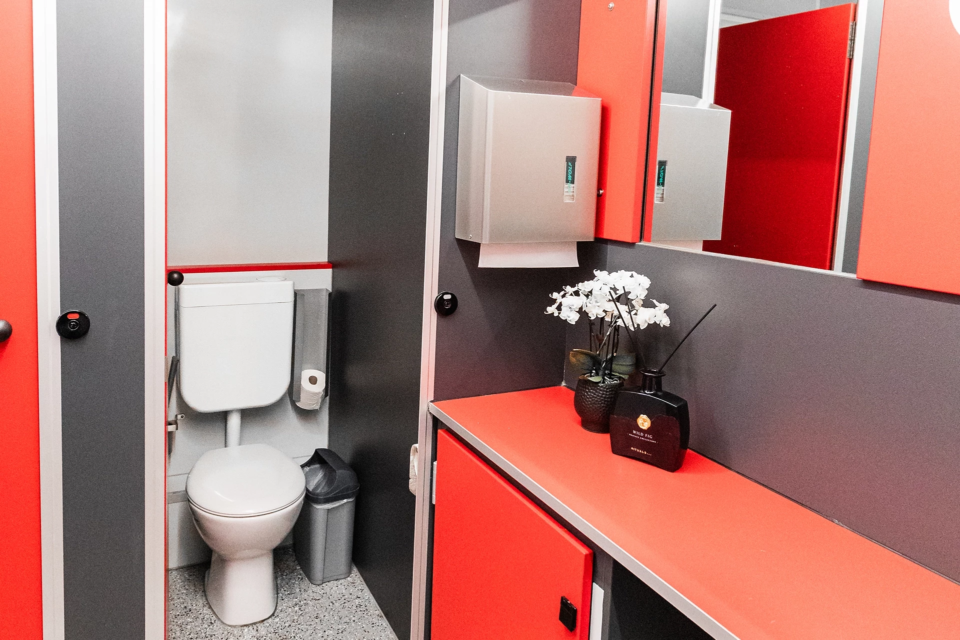 Toilettenwagen Modell Exklusiv – Mix 3/2 10 Rotes und graues Badezimmer mit Toilette, Waschbecken, Papiertuchspender und weißen Blumen in einer Vase.