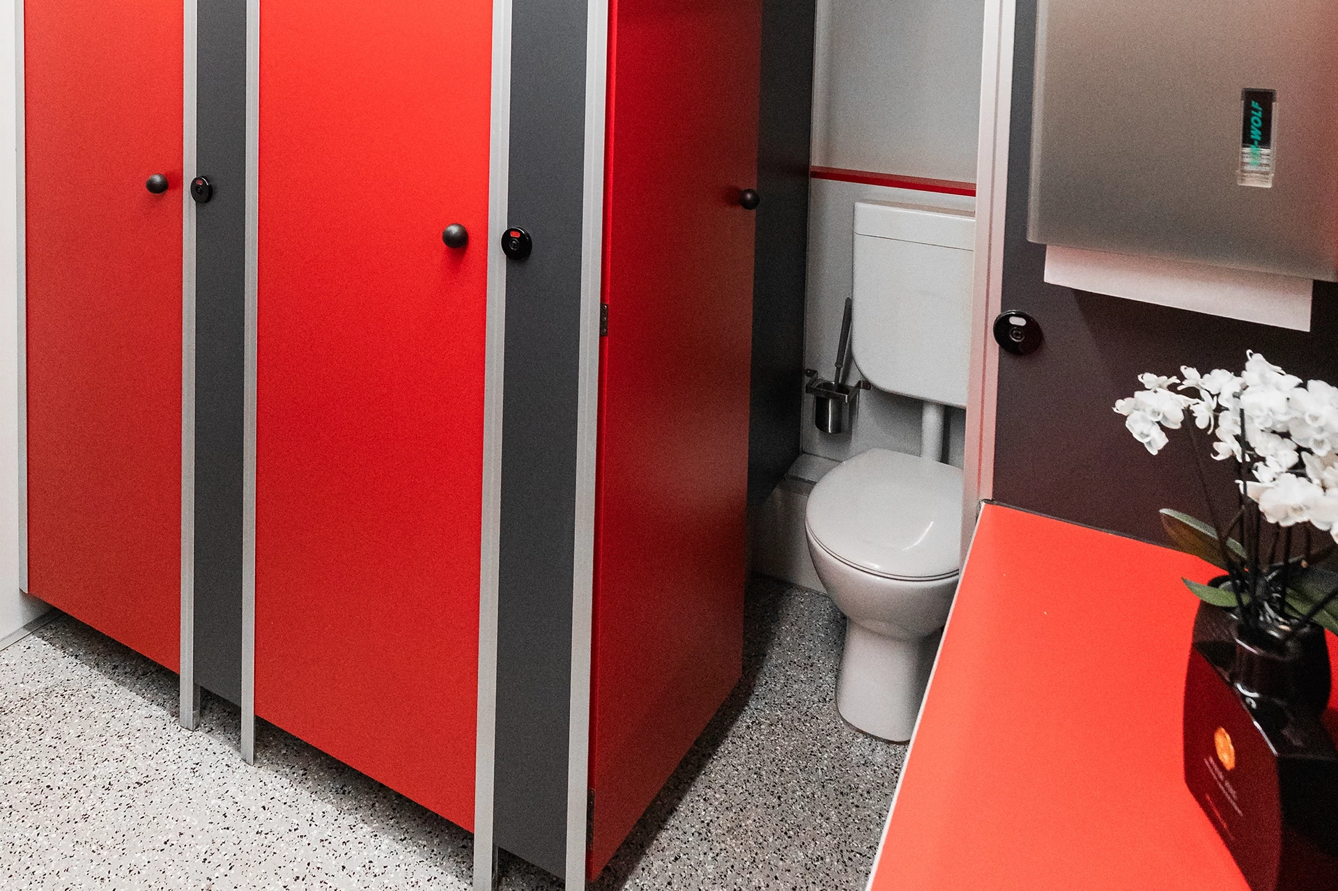 Toilettenwagen Modell Exklusiv – Mix 3/2 8 Öffentliche Toilette mit roten Kabinentüren, grauem Boden und einer Vase mit weißen Blumen auf einer Ablage.