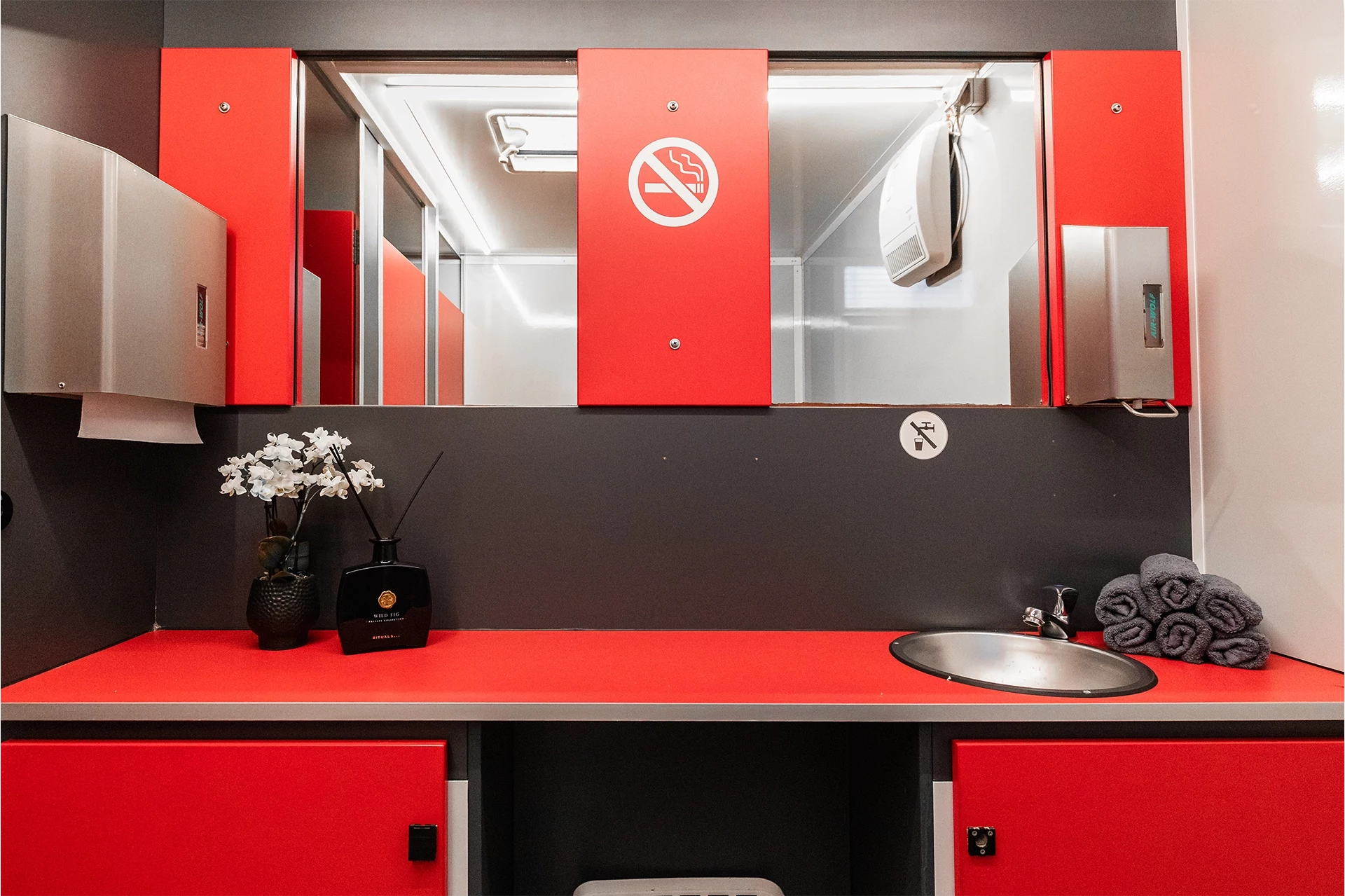 Toilettenwagen Modell Exklusiv – Mix 3/2 6 Moderne Toilette mit rotem Waschbecken, Spiegel, Handtuchrollen, Orchidee und Nichtraucher-Schild an der Trennwand.