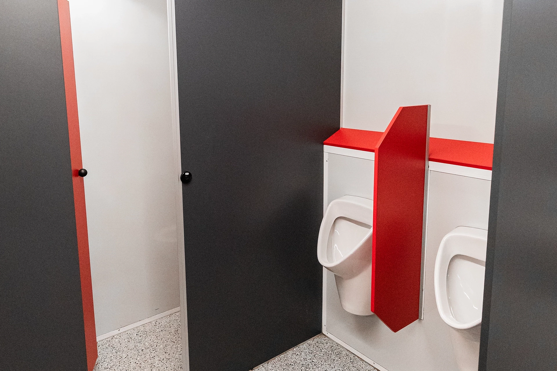 Toilettenwagen Modell Exklusiv – Mix 3/2 4 Zwei Urinale mit rotem Trennwand in einer grau-weißen Toilette mit geschlossenen Kabinentüren und grauem Boden.