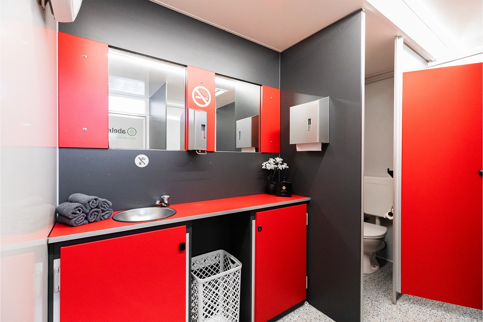 Toilettenwagen Modell Exklusiv – Mix 3/2 3 Moderne öffentliche Toilette mit rotem und grauem Design, Waschbecken, Spiegel, Handtuchspender und offener Kabine.