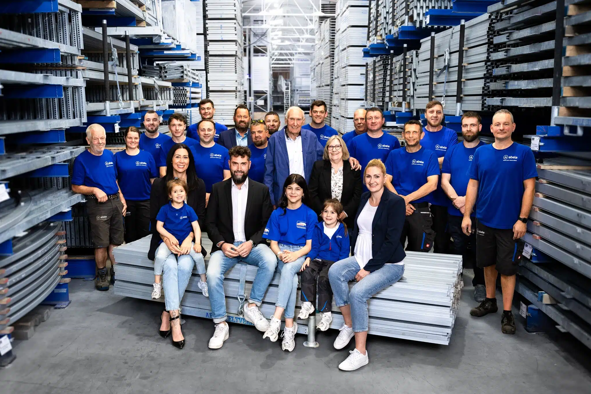 Gruppenfoto eines Teams in blauen Shirts, das in einem Lager auf Metallstangen sitzt und steht, ausgleuchtet von oben.