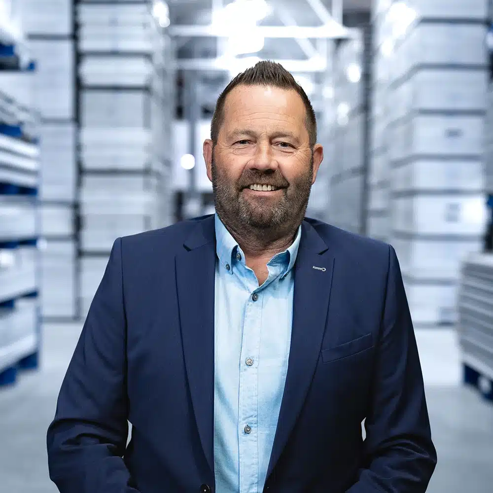 Mann in Anzug lächelt in Lagerhalle, umgeben von hohen Regalen voller gestapelter Kisten.