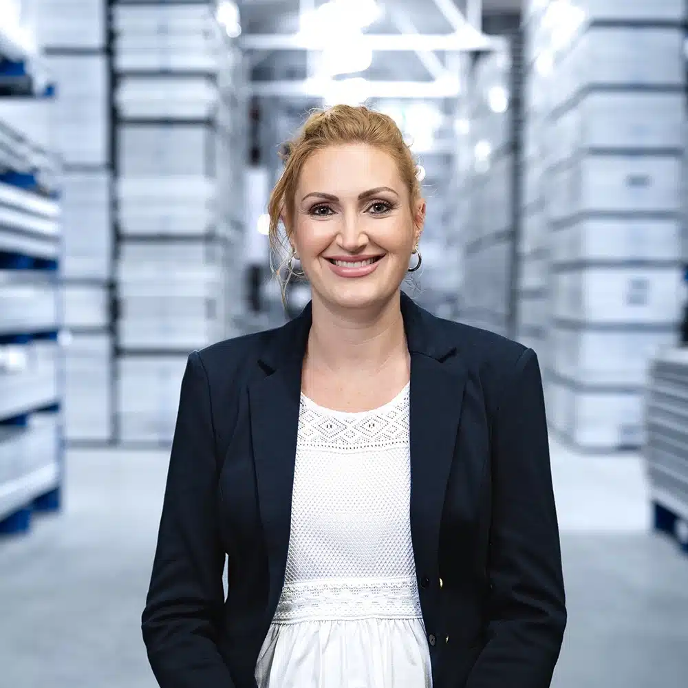Eine lächelnde Frau in Blazer steht in einem Lager mit gestapelten Kisten im Hintergrund.