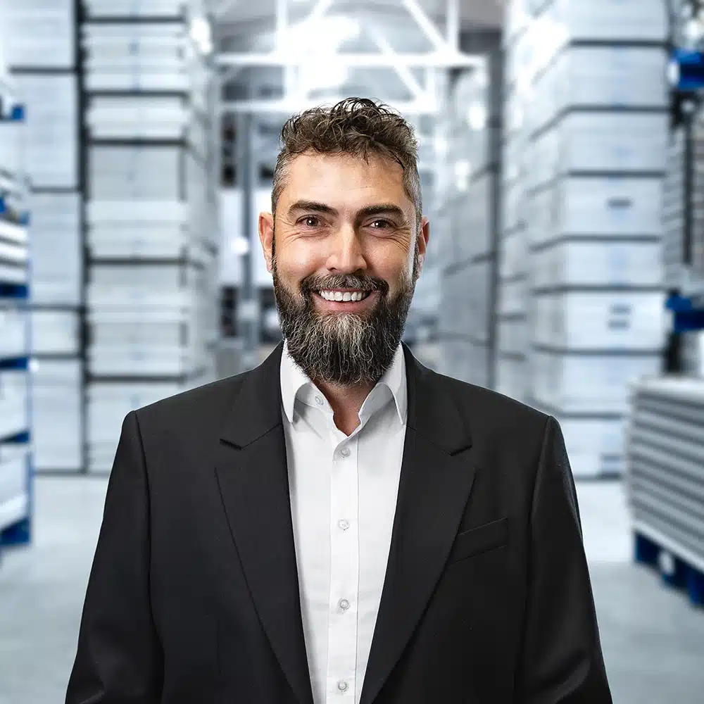 Mann mit Bart im Anzug lächelt in einem Lagerraum mit hohen Regalstapeln im Hintergrund.