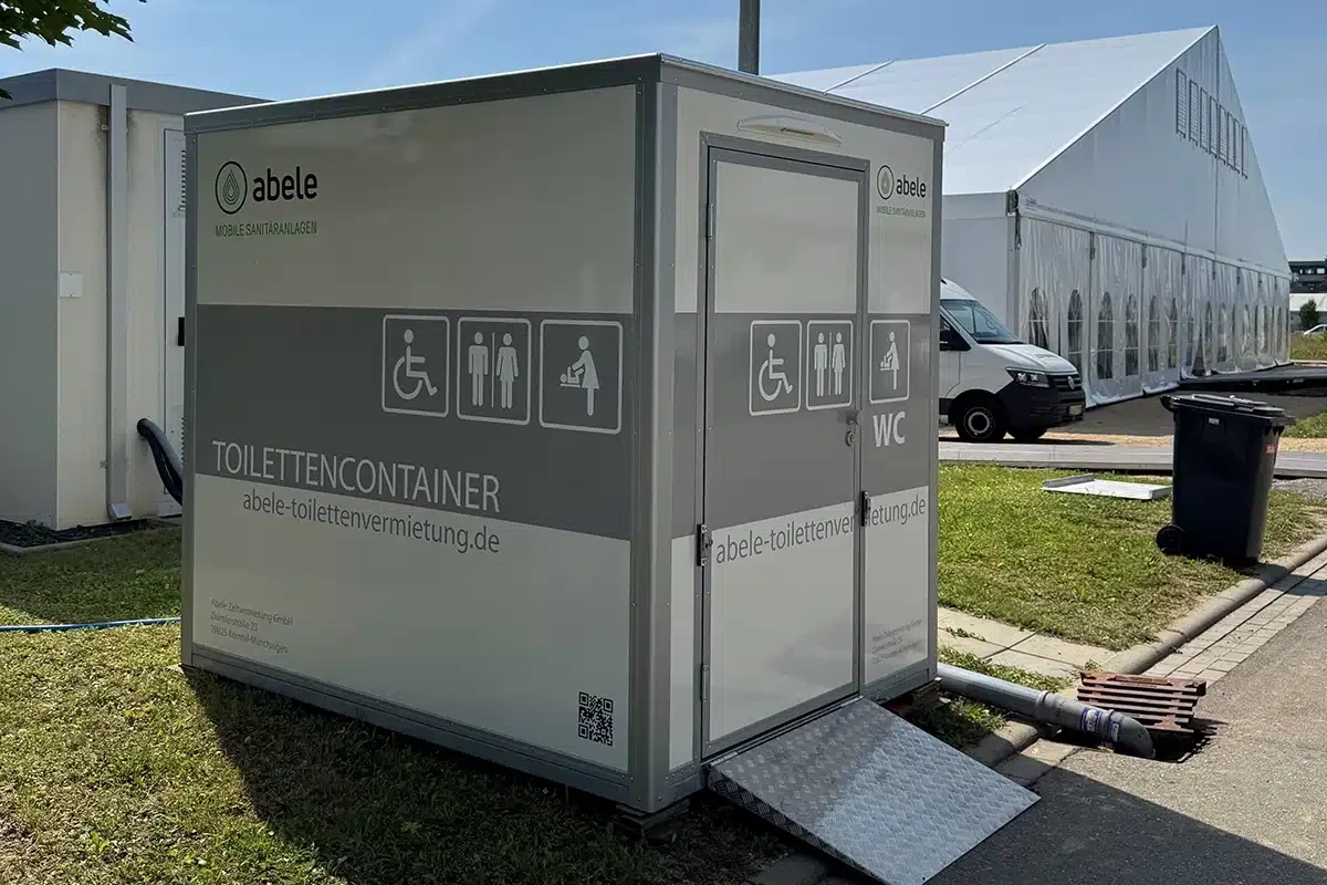 Toilettenvermietung Stuttgart 2 Mobiler Toilettencontainer mit Rollstuhlzugang, Symbolen für Barrierefreiheit und Babywickeltisch, neben einem Zelt und Van.