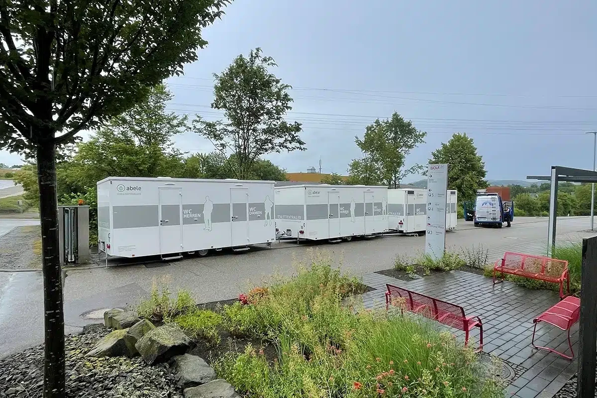 Toilettenvermietung Stuttgart 8 Mobile Toilettenanhänger und ein Servicefahrzeug stehen auf einem Parkplatz, umgeben von Bäumen und Sitzbänken.