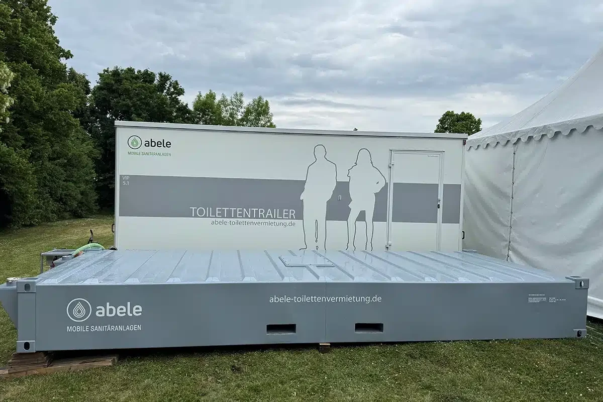 Toilettenvermietung Stuttgart 10 Mobiler Toiletten-Trailer auf grünem Rasen vor Bäumen und einem Zelt, mit Firmenlogo und stilisierter Personen-Silhouette.