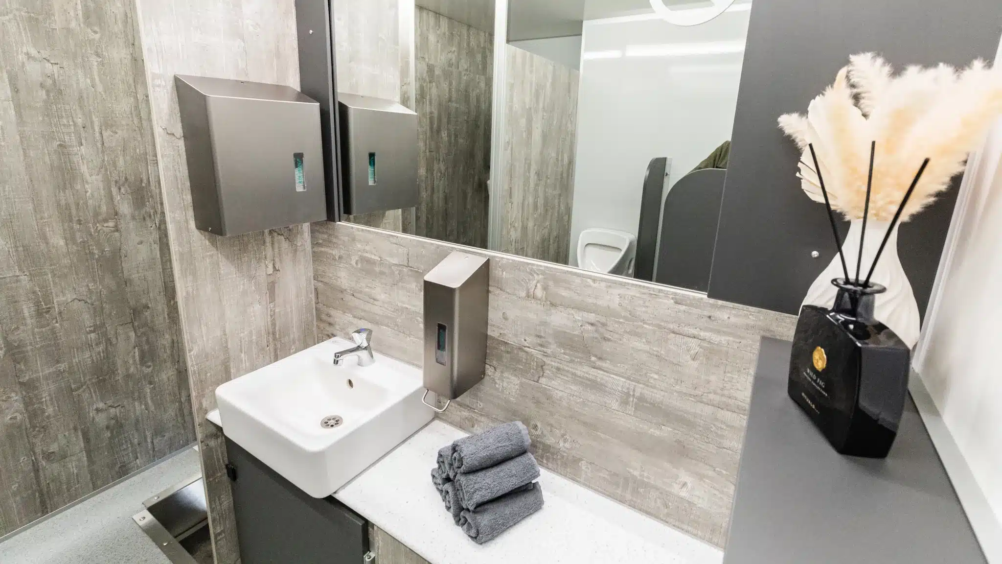 Unterseite Toilettenwagen „Standorte“ Moderner Badezimmerbereich mit Waschbecken, Spiegel, Seifenspender, Handtüchern und Dekoration vor Holzwand.