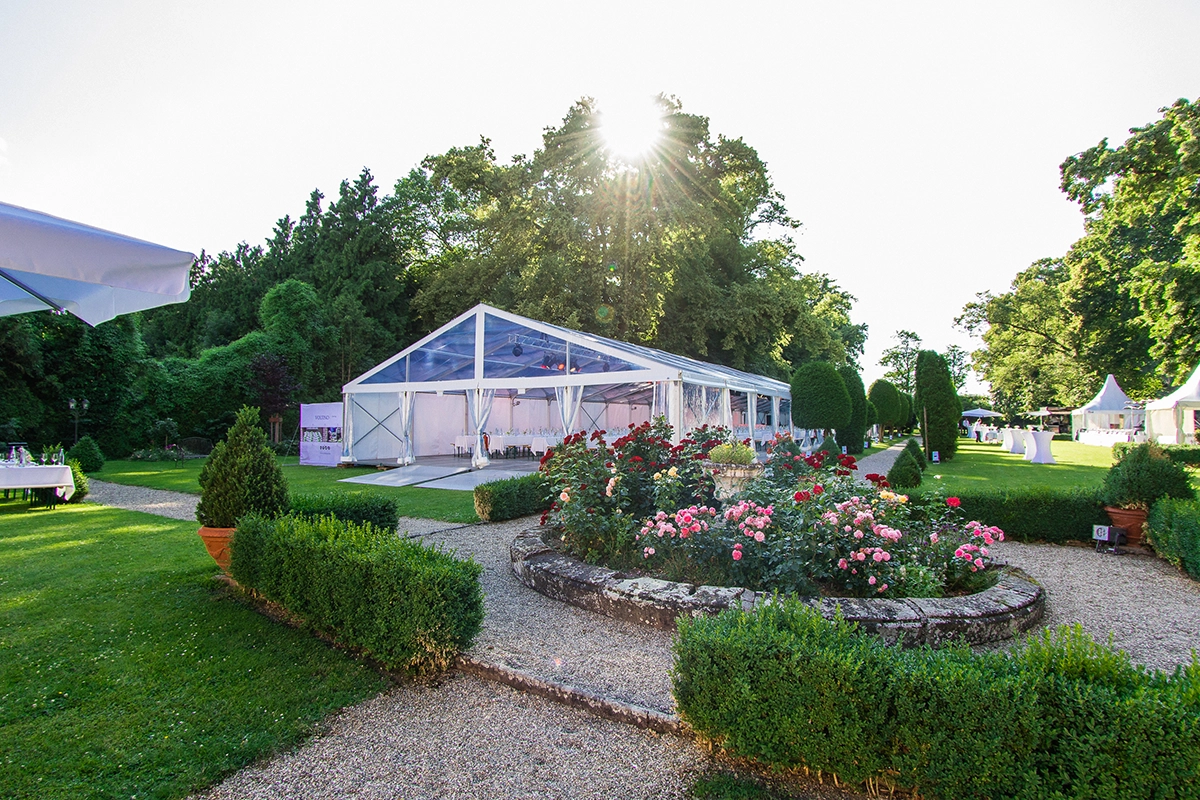 Großes, transparentes Festzelt in einem Garten mit blühenden Rosen und sonnigem Himmel im Hintergrund.