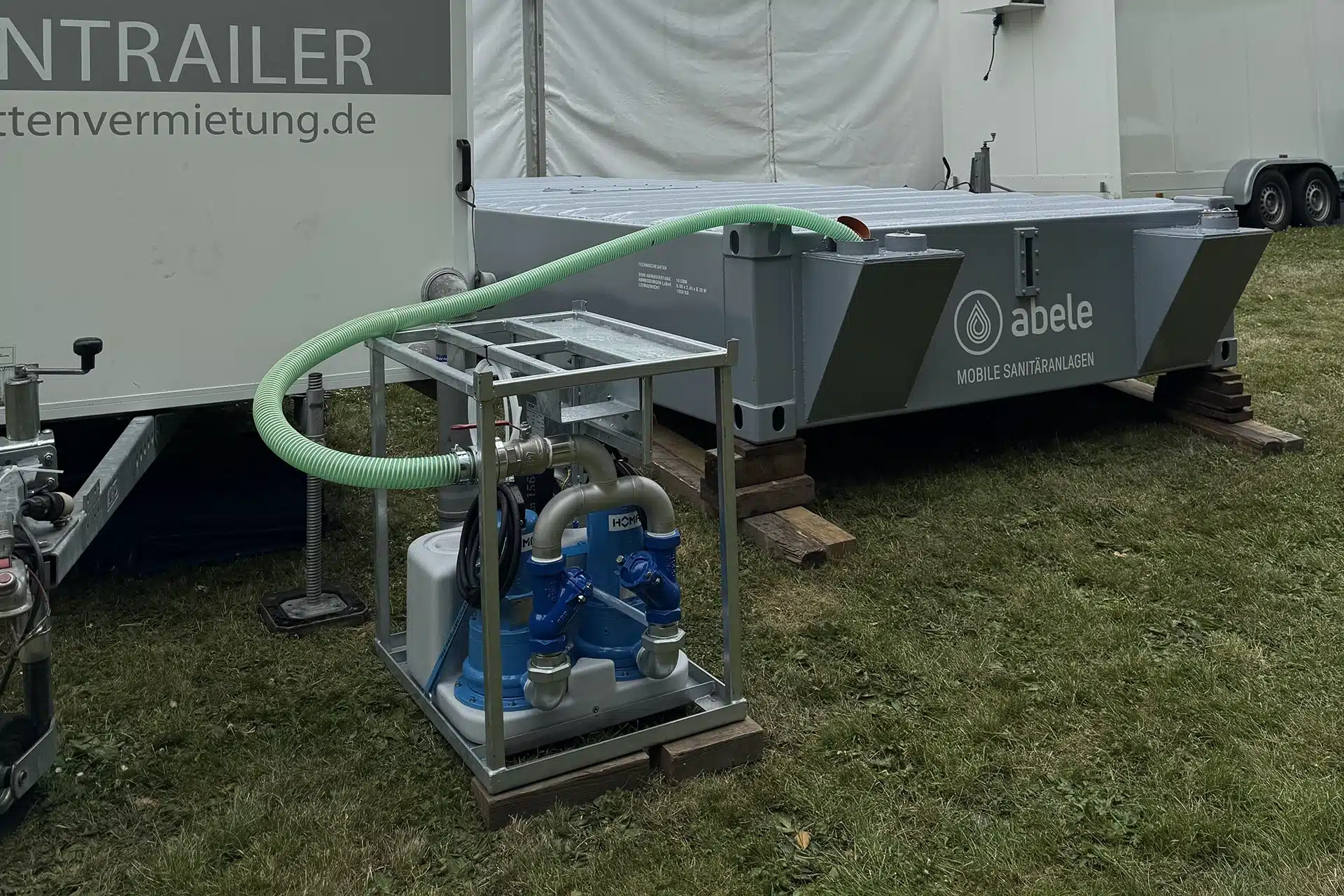 Abwassertank / Pumpe 7 Mobile Sanitäranlage mit Rohren und Pumpe auf Wiese, im Hintergrund ein Anhänger mit Werbeaufschrift.