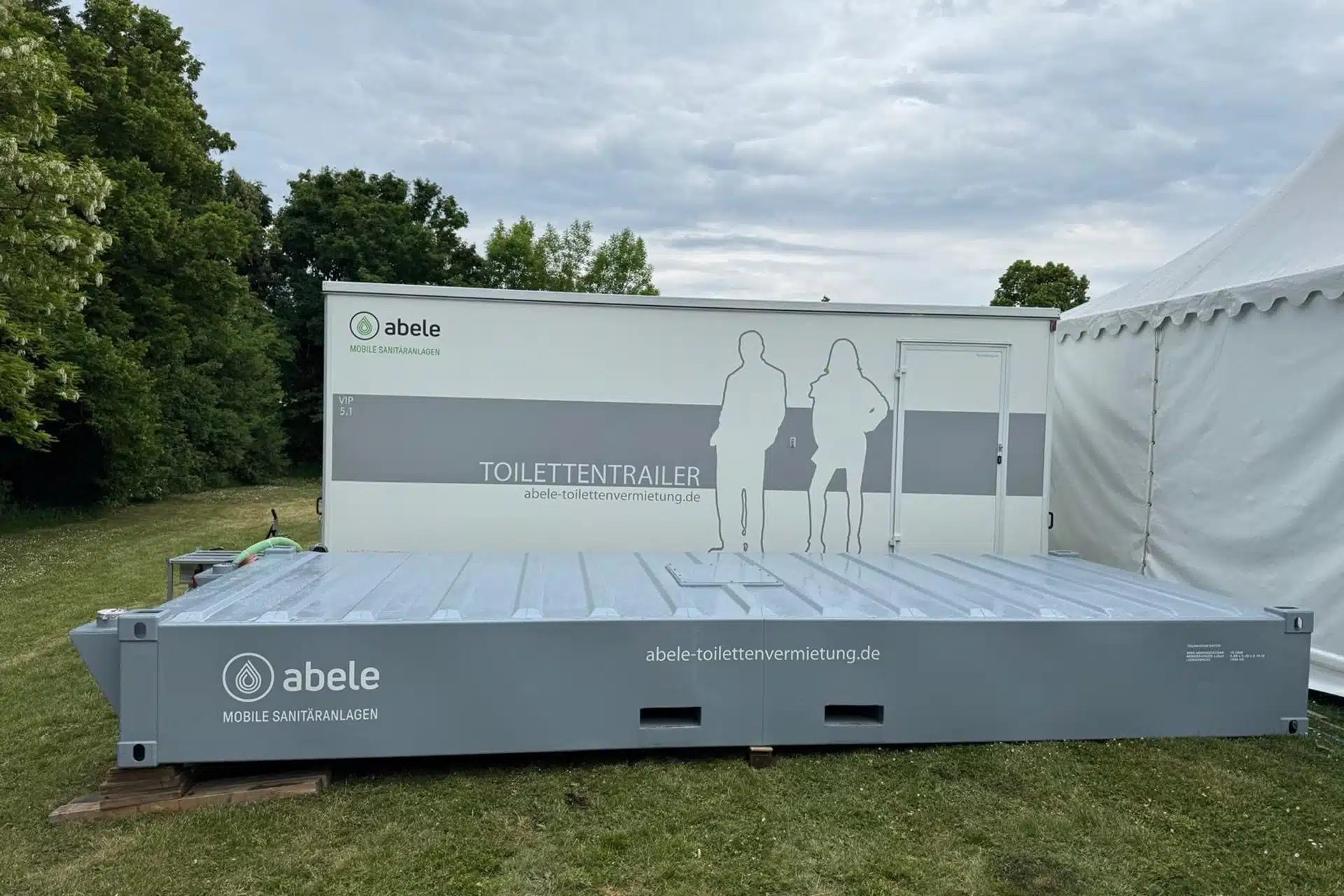 Abwassertank / Pumpe 9 Mobiler Toilettentrailer mit grauem Design, umgeben von Grünflächen und einem Zelt im Hintergrund.